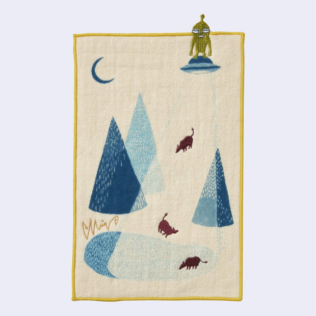 Rectangular cream colored towel with a yellow border, of several cows amongst a lake and pointed mountains, being abducted but a UFO with a small green alien standing on top..
