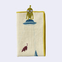 Rectangular cream colored towel with a yellow border, of several cows amongst a lake and pointed mountains, being abducted but a UFO with a small green alien standing on top. Folded.