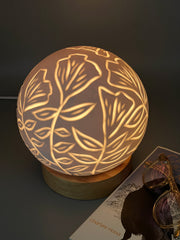 Spherical lamp with carved imagery of poppy flowers and leaves. It is lit and soft light comes out from the flower shapes.