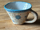 Ceramic mug, mostly cream colored with a blue rim and design of a blue starburst with red lines on the back of the mug.