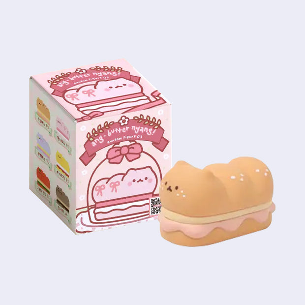 Small cartoon style toy of a cat sandwich cake, with its body like a long loaf. It sits in front of its product packaging. 