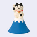 Cat figurine posed like a lucky cat, with one paw up, sitting atop of Mt. Fuji, blue with a white capping of snow.