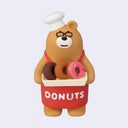 Figurine of a brown bear wearing glasses, a chef's hat and a red apron that reads "donuts". It holds a tray of donuts.
