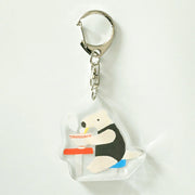 Acrylic keychain of an anteater sitting at a stool and eating a steaming bowl of noodles.