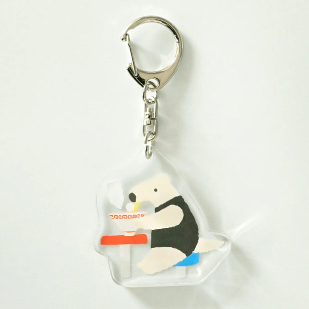 Acrylic keychain of an anteater sitting at a stool and eating a steaming bowl of noodles.