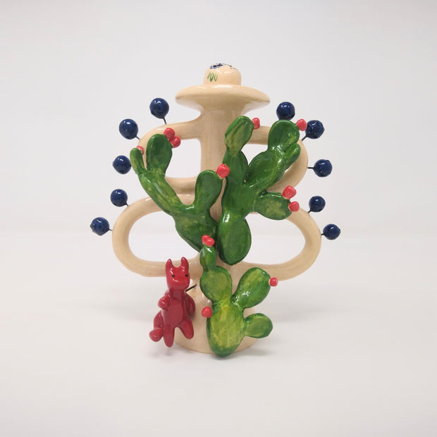 Ceramic sculpture of tree made out of cacti, with flowering red buds. A small red devil sits at the base, and the whole tree is attached to a cream colored stand with loops and blue dots extended out.
