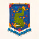 Decorative wall hanging with a green alligator design on a blue background, featuring colorful stars and pom-poms.