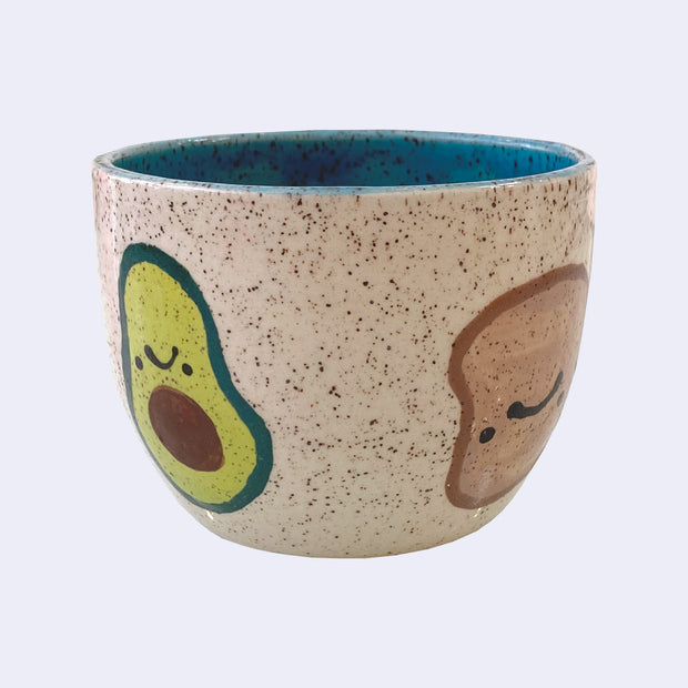 Ceramic bowl with spotted finishing and an earthy brown exterior and teal blue interior. On the outside are painted on cartoon style avocado and toast, with simple expressions.