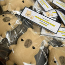 A pile of plush capybara toys in clear plastic bags with printed tags. The toys are a light tan/beige color with embroidered black eyes and a darker brown stitched mouth area.