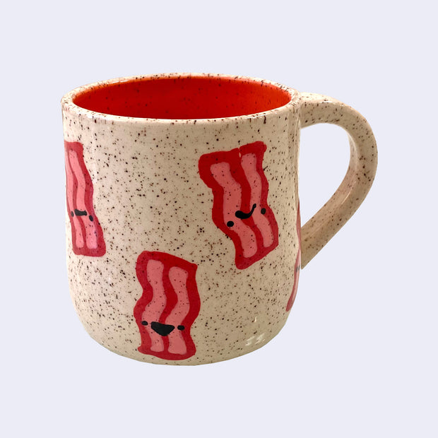 Ceramic mug with spotted finishing and an earthy brown exterior and pinkish red interior. On the outside are painted on cartoon style bacon, with simple expressions.
