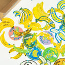 Scattered array of banana themed flake stickers, all with a slight holographic shine. Bananas are either realistic depictions, cartoons, or look like fruit label stickers.