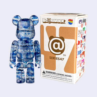 Vinyl bear figure with blocky body features, like an action figure. It has no facial features and its whole body is covered in a shiny blue and silver futuristic sparkle pattern. It stands next to its product packaging.