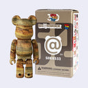 A Bearbrick Series 52 collectible figure with a brown and beige wood-grain patterned body, standing next to its original packaging box on a light gray background.