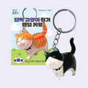 Keychain of a smiling black tuxedo cat, with a white stomach and part of its face. It is in mid-stride with a curved, upward tail. Its attached to a keyring and stands in front of its product packaging.