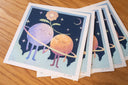 Stack of prints featuring an illlustration of 2 anthropomorphized moons, holding hands with a flower aboved their heads. They stand within a ring against a starry night sky