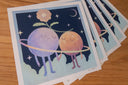 Stack of prints featuring an illlustration of 2 anthropomorphized moons, holding hands with a flower aboved their heads. They stand within a ring against a starry night sky