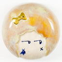 Round ceramic tile of a head with a round, puffy hair with a gold hair bow and a face with watchful eyes and "x" on cheeks.