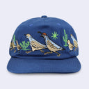 A blue baseball cap featuring detailed embroidery of quail birds, cacti, and a green star on the front panel, set against a light gray background.