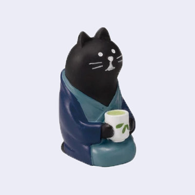 Small figure of a black cat wearing a blue kimono and sitting on the ground while holding a cup of green tea.
