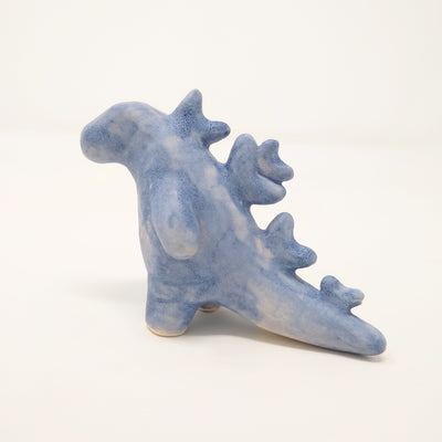 Ceramic sculpture of Godzilla, made of mostly smooth shapes and features. It has stylized spikes along its back and no rendered facial features. Colors are a matte purplish blue.