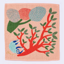 Salmon pink square towel with illustration of a large, red tree with green leaves, hanging off of it is a fluffy blue sloth. Next to it is another stylized tree in a similar color scheme. 