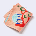 Salmon pink square towel with illustration of a large, red tree with green leaves, hanging off of it is a fluffy blue sloth. Next to it is another stylized tree in a similar color scheme. 