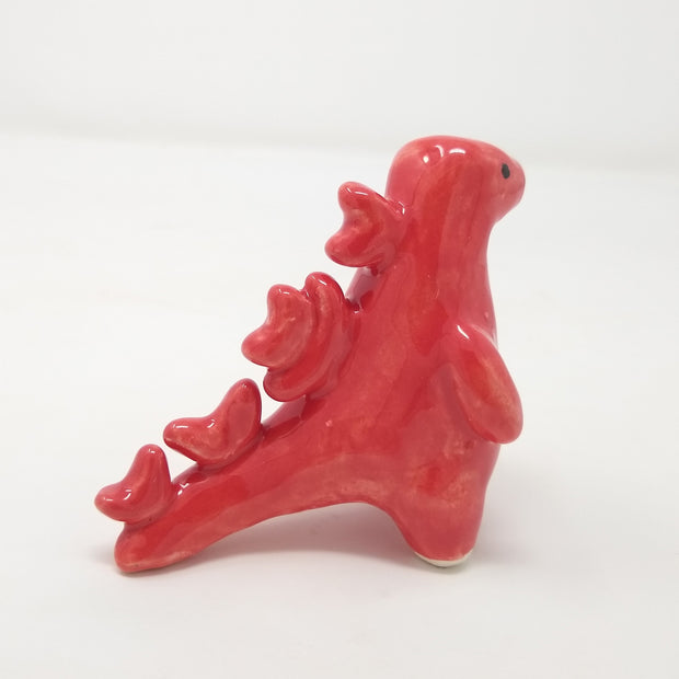 Ceramic sculpture of Godzilla, made of smooth shapes and features. It has stylized spikes on its back and a set of rendered eyes on its otherwise plain face. Colors are glossy reddish pink.