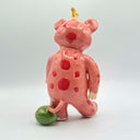 Glazed ceramic sculpture of a pink bear-like monster with cute, buck-teeth and a yellow crown of sorts. Its body has pink polka dots and it balances its tail on a small watermelon.