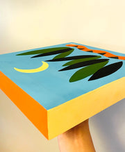 Solid color painting on panel of a blue background. A simply painted orange and black snake is at the bottom between blades of tall grass. A simplistic crescent moon hangs overhead.