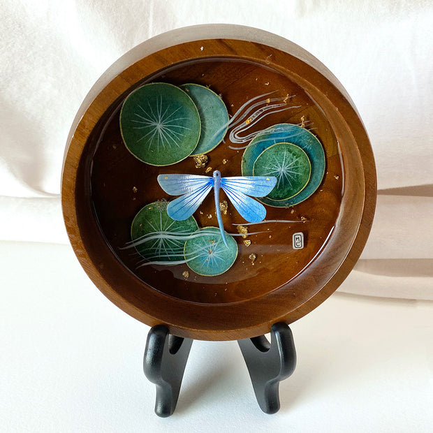 Flat bottomed dark grain wooden bowl containing a scene of painted cut paper, sealed in with resin. A blue dragonfly rests among various round lily pads, with gold flecks throughout. 