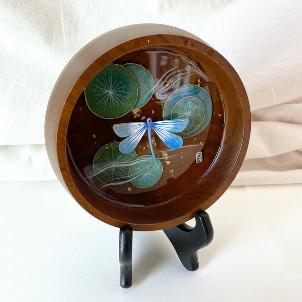 Flat bottomed dark grain wooden bowl containing a scene of painted cut paper, sealed in with resin. A blue dragonfly rests among various round lily pads, with gold flecks throughout. Displayed from the side.