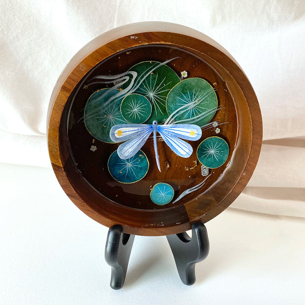 Flat bottomed dark grain wooden bowl containing a scene of painted cut paper, sealed in with resin. A indigo blue dragonfly rests among various round lily pads, with gold flecks throughout.