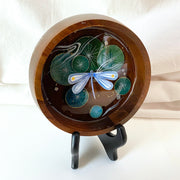 Flat bottomed dark grain wooden bowl containing a scene of painted cut paper, sealed in with resin. A blue dragonfly rests among various round lily pads, with gold flecks and small white flowers throughout.