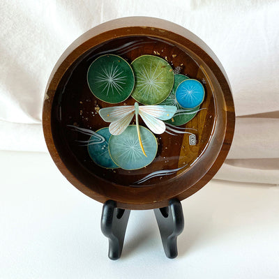 Flat bottomed dark grain wooden bowl containing a scene of painted cut paper, sealed in with resin. A white and mint colored dragonfly rests among various round lily pads, with gold flecks throughout.