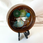 Flat bottomed dark grain wooden bowl containing a scene of painted cut paper, sealed in with resin. A white and mint colored dragonfly rests among various round lily pads, with gold flecks throughout.