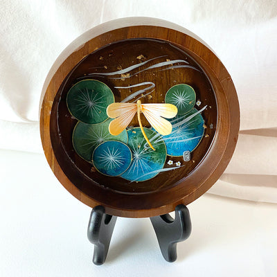 Flat bottomed dark grain wooden bowl containing a scene of painted cut paper, sealed in with resin. A yellow dragonfly rests among various round lily pads, with gold flecks and small white flowers throughout.