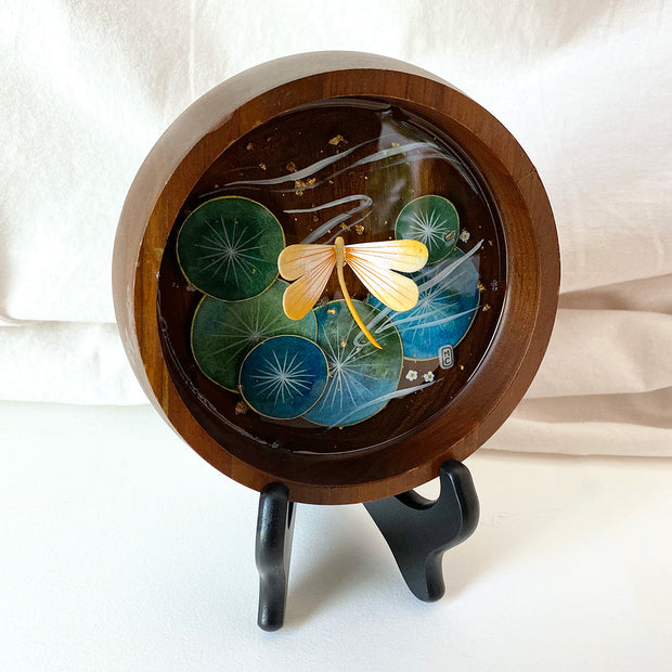 Flat bottomed dark grain wooden bowl containing a scene of painted cut paper, sealed in with resin. A yellow dragonfly rests among various round lily pads, with gold flecks and small white flowers throughout.