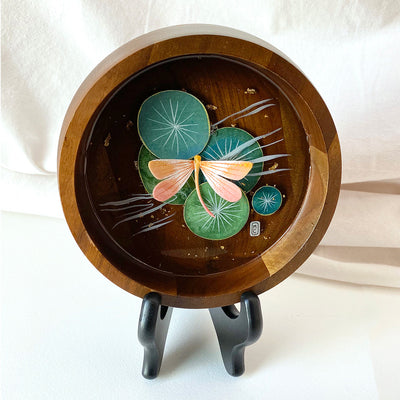 Flat bottomed dark grain wooden bowl containing a scene of painted cut paper, sealed in with resin. A peach colored dragonfly rests among various round lily pads, with gold flecks throughout.