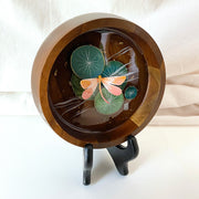 Flat bottomed dark grain wooden bowl containing a scene of painted cut paper, sealed in with resin. A peach colored dragonfly rests among various round lily pads, with gold flecks throughout.