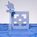 Carved wooden sculpture made to look like a cement breeze block. The block is entirely painted a light purplish blue and a tall sign post stands nearby, painted in similar colors.