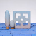 Carved wooden sculpture made to look like a cement breeze block. The block is entirely painted a light blue to pink gradient. Two oval shaped trees of varying heights stand nearby, painted in similar color tones.