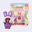 A Calico Critters collectible bunny figure in a purple polka-dot outfit with a white hood stands in front of its retail packaging on a light gray background.