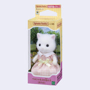 White cat figurine with flocked fur, wearing a pink checkered dress and yellow bow. It is in its product packaging box.