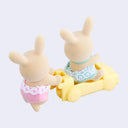 Two small rabbit dolls with cream colored flocked fur, dressed in small children's clothing. One pushes the other in a yellow car.