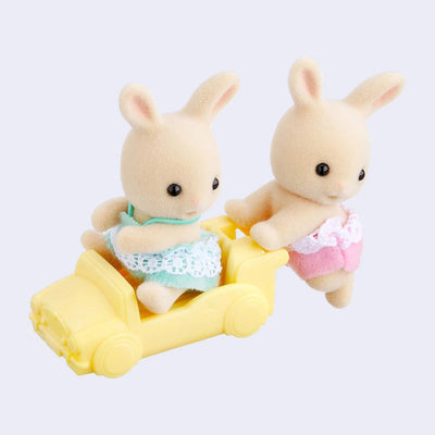 Two small rabbit dolls with cream colored flocked fur, dressed in small children's clothing. One pushes the other in a yellow car.
