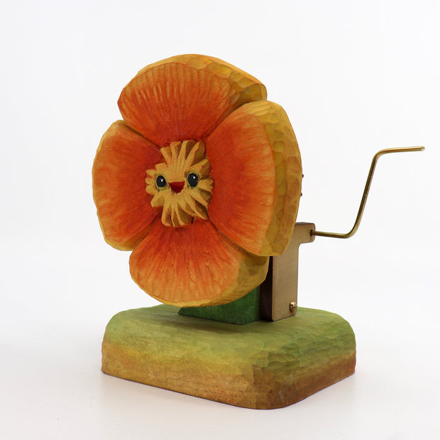 Small wooden sculpture of a orange poppy with a small cartoonish face. Sculpture is on a stand and has a lever attached to the back.