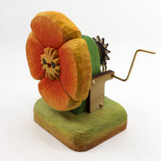 Small wooden sculpture of a orange poppy with a small cartoonish face. Sculpture is on a stand and has a lever attached to the back.