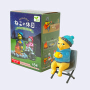 Small plastic yellow cat dressed in a warm blue jacket and knit cap, sitting in a camping chair and holding a yellow mug. It sits in front of its product packaging.