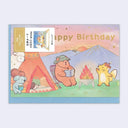 A Japanese Happy Birthday greeting card featuring cute cartoon animals camping outdoors. The illustration shows a bear reading by a campfire next to a llama and a small creature by a tent, with a mountain and sunset in the background.