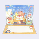 A 3D pop-up greeting card featuring cute animal characters celebrating with a cake and campfire. The card is shown partially unfolded to reveal the bottom panel with Japanese text and small illustrations, set against a plain light gray background.
